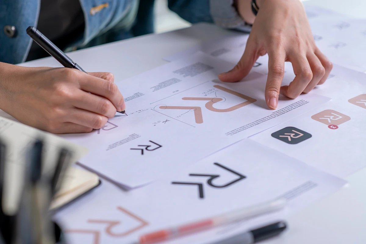 Designer sketching brand logo concepts on printed guides, analyzing geometry and icon variations during brand identity development.