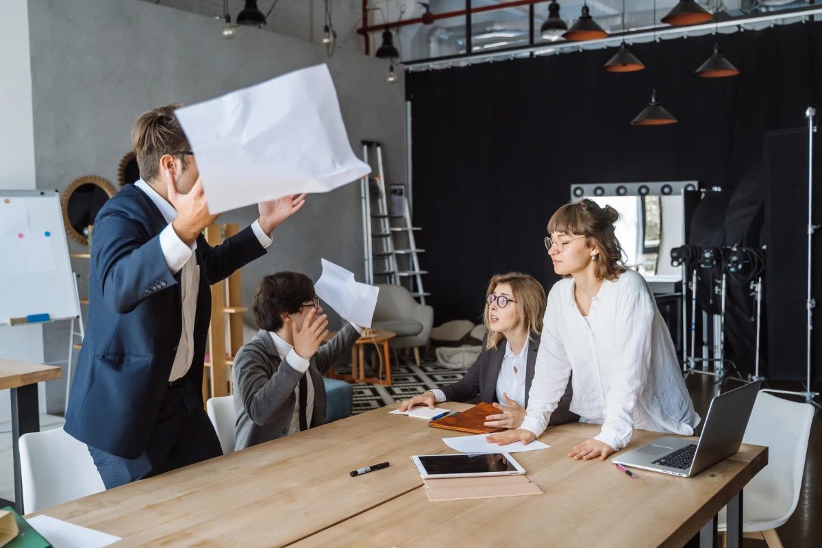 Creative team in a heated design discussion, one member standing while another throws papers in frustration.