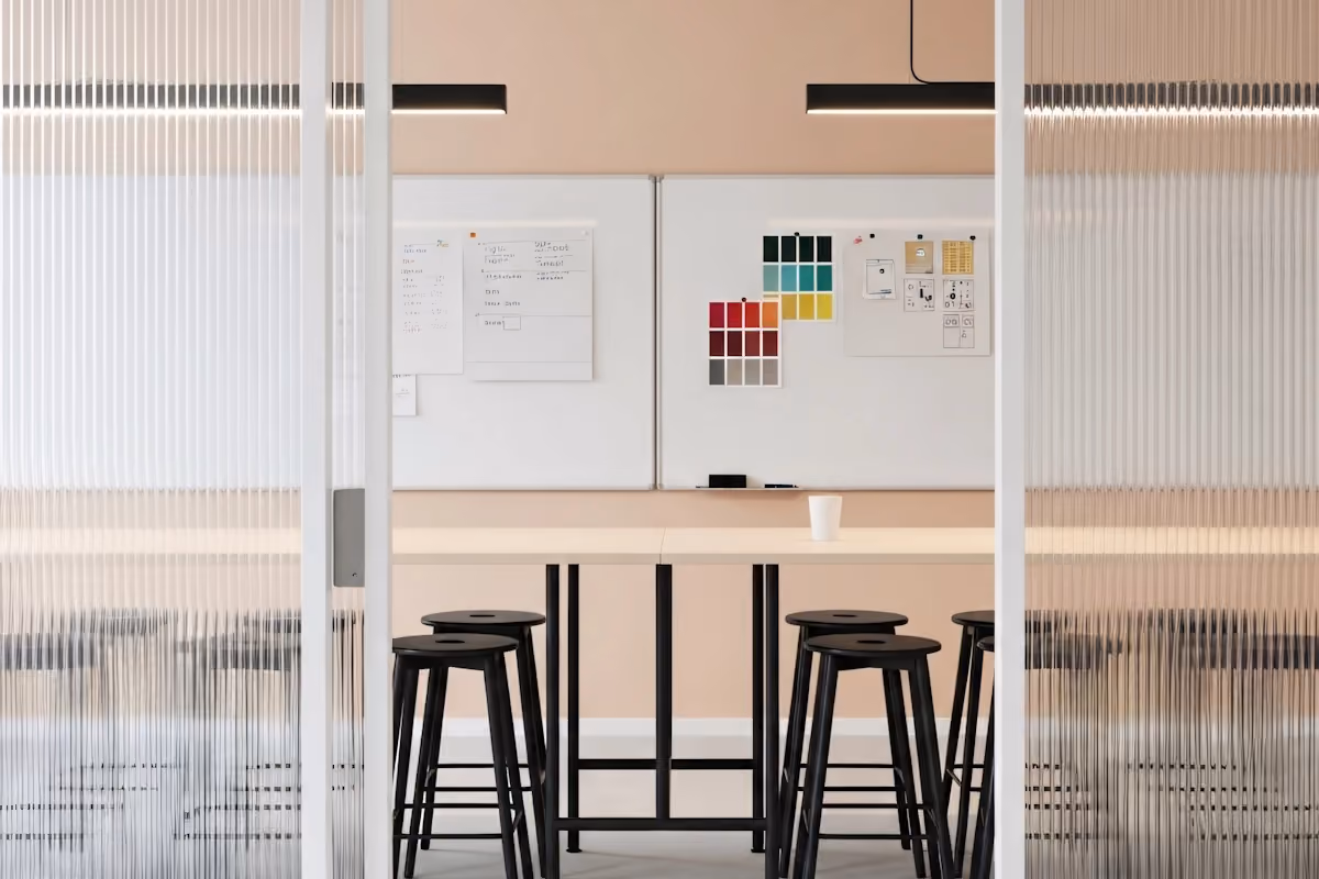 A design meeting space with high stools, a whiteboard filled with color palettes and UI sketches, and soft lighting viewed through a ribbed glass partition.