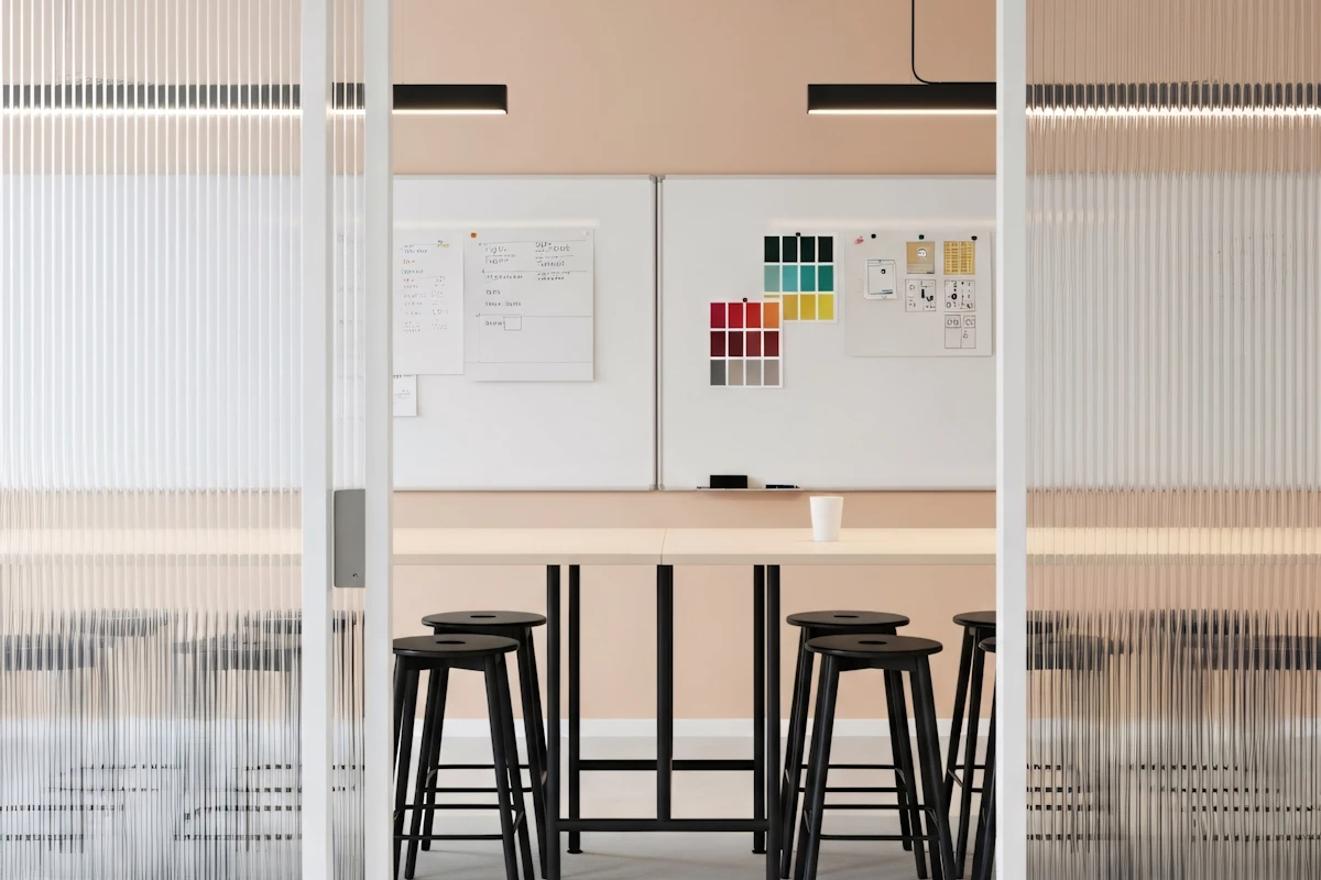A design meeting space with high stools, a whiteboard filled with color palettes and UI sketches, and soft lighting viewed through a ribbed glass partition.