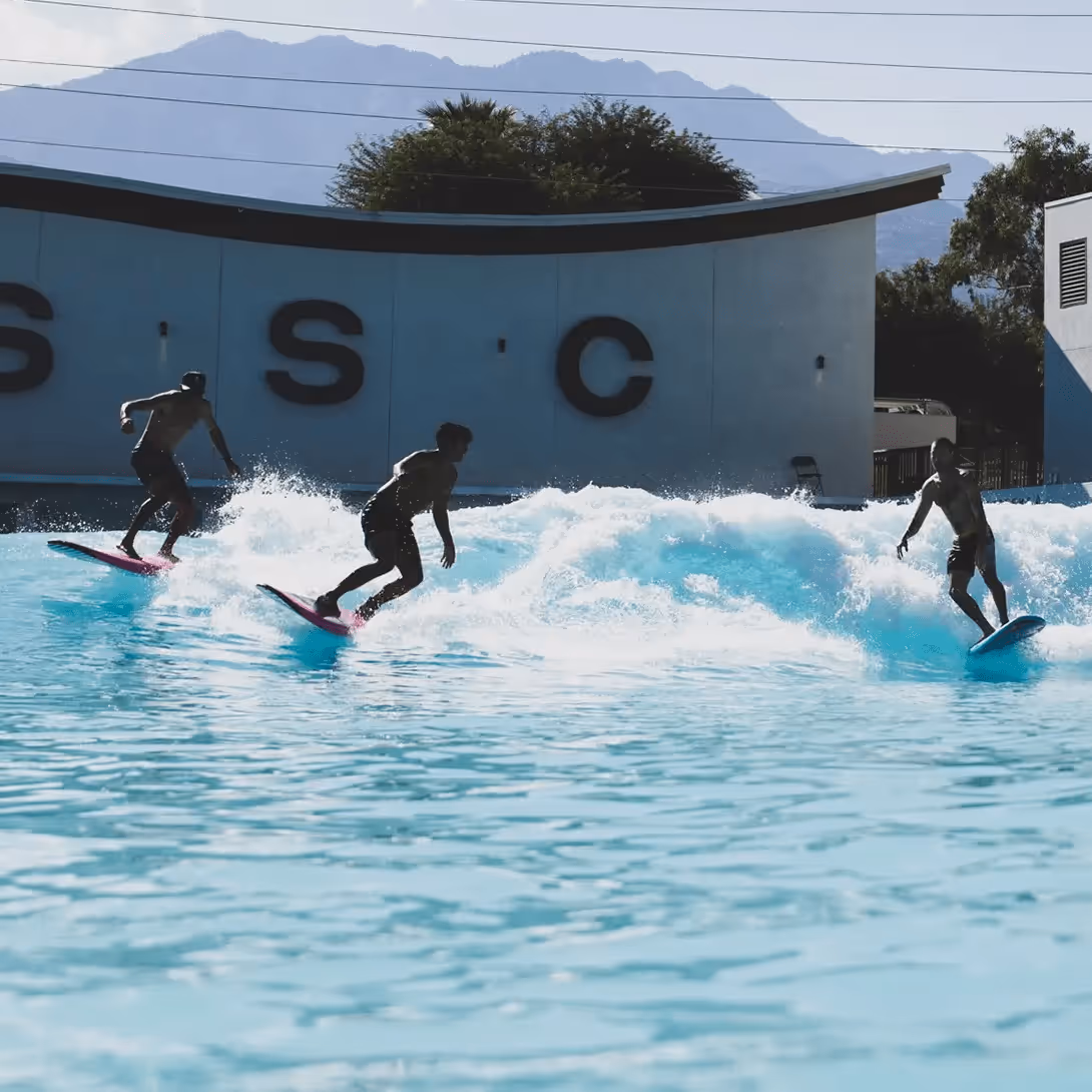 Three surfers riding an artificial wave at Palm Springs Surf Club, with turquoise water splashing around them and the letters “PSSC” visible on the wall in the background.