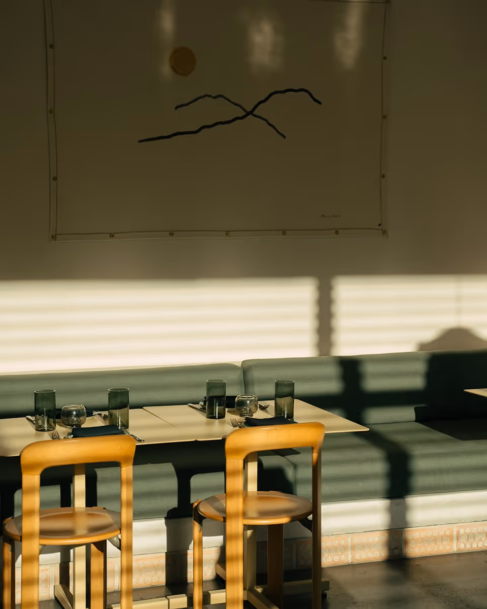 Minimalist restaurant interior with green upholstered bench seating, wooden chairs, and neatly set tables, bathed in warm sunlight casting shadows from window blinds.