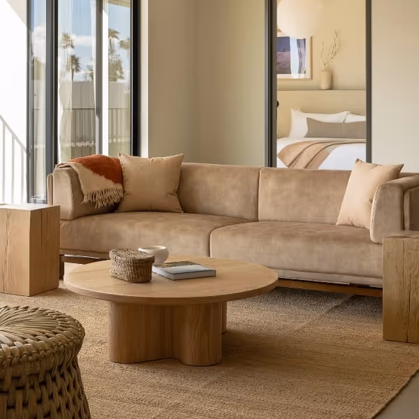 Cozy neutral-toned living room with a tan sectional sofa, round wooden coffee table, woven ottoman, and a view into a neatly styled bedroom.