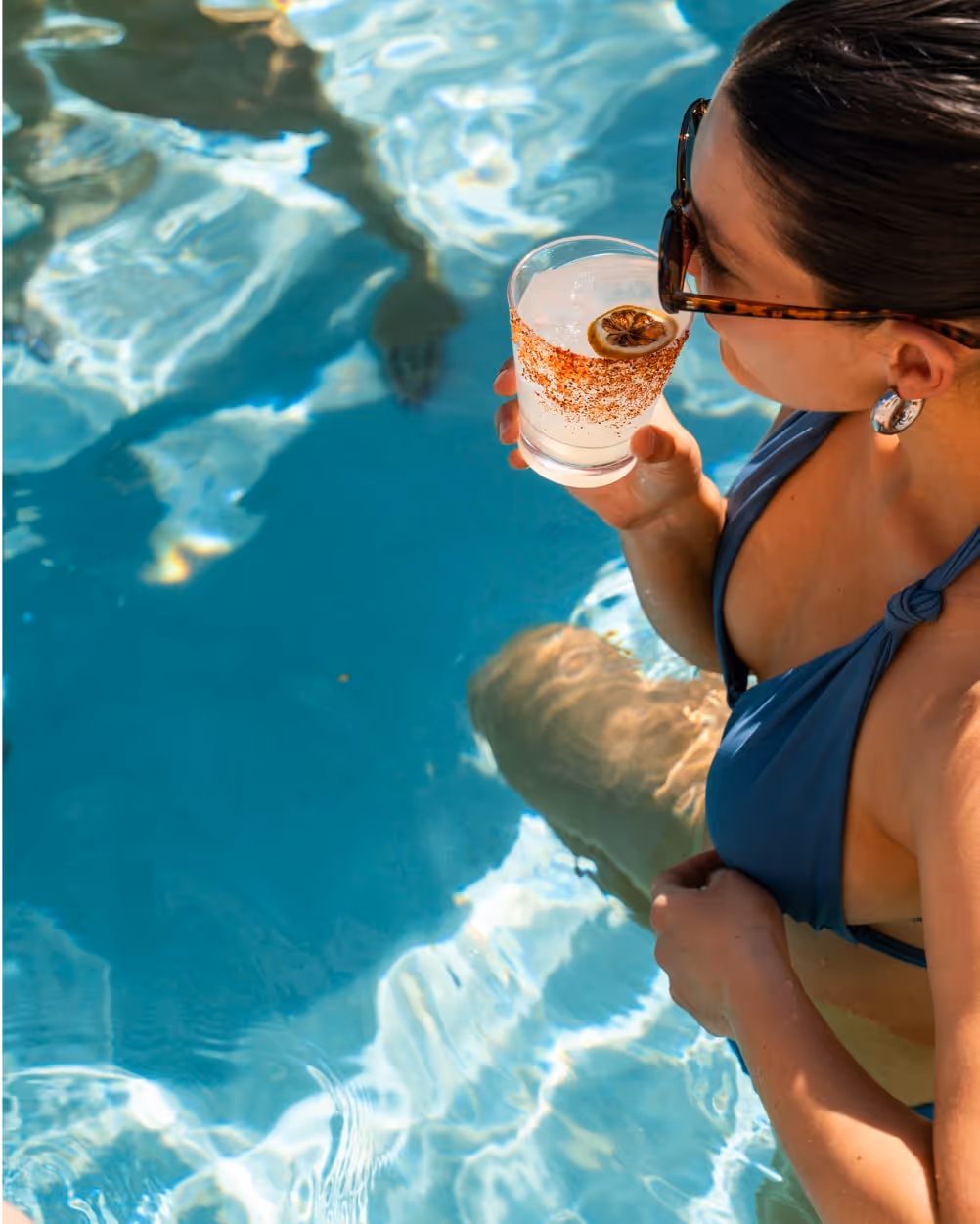 Woman in a blue bikini enjoying a cocktail with a dried lime garnish while relaxing in a swimming pool under the sun.