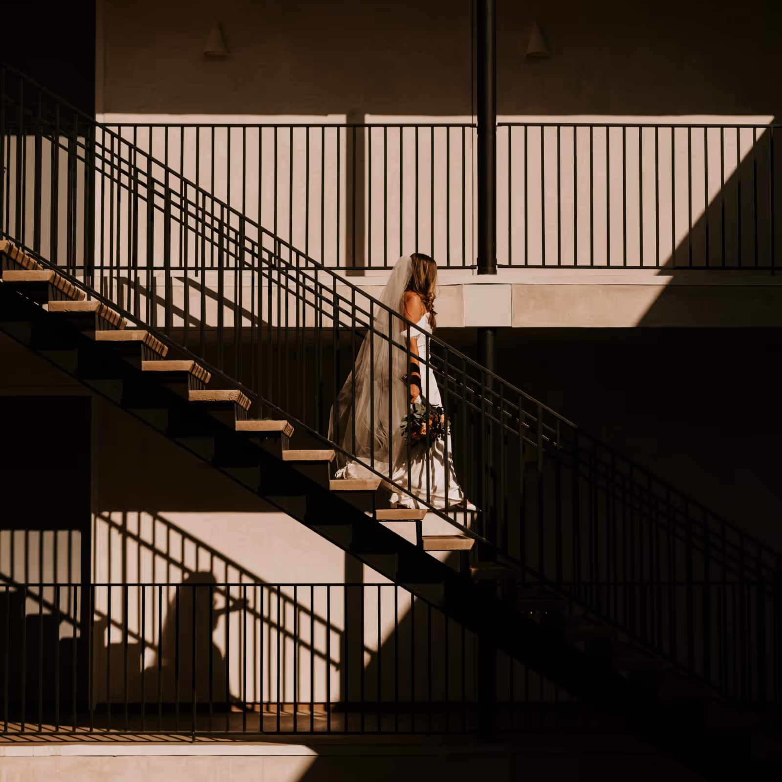Bride in a white gown and veil holding a bouquet, walking up a sunlit staircase casting dramatic shadows on a modern building.