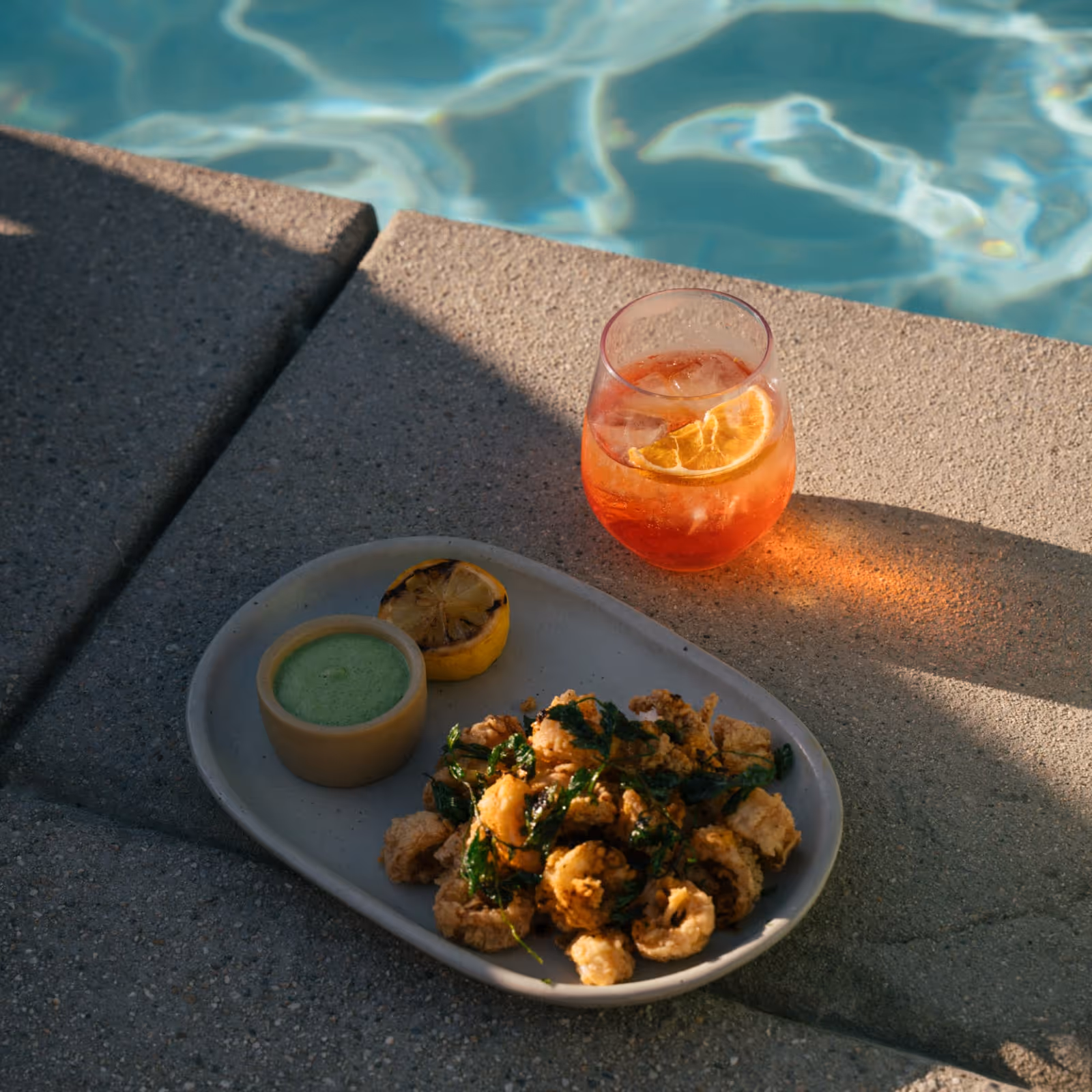 Close-up of a poolside snack featuring crispy fried calamari with dipping sauce, grilled lemon, and a cocktail garnished with dried citrus slices.