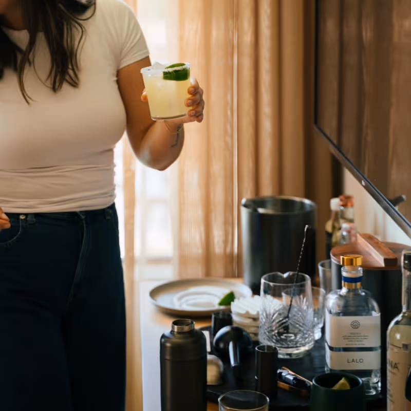 Woman holding a margarita garnished with lime, standing at a bar setup with cocktail tools, tequila bottles, and ingredients on a table.