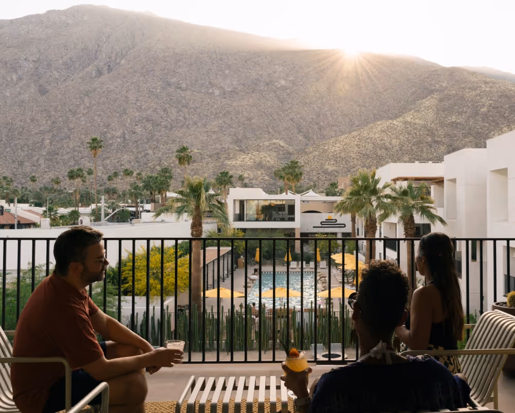 Three people enjoy drinks on a balcony overlooking a resort pool, palm trees, and desert mountains at sunset.