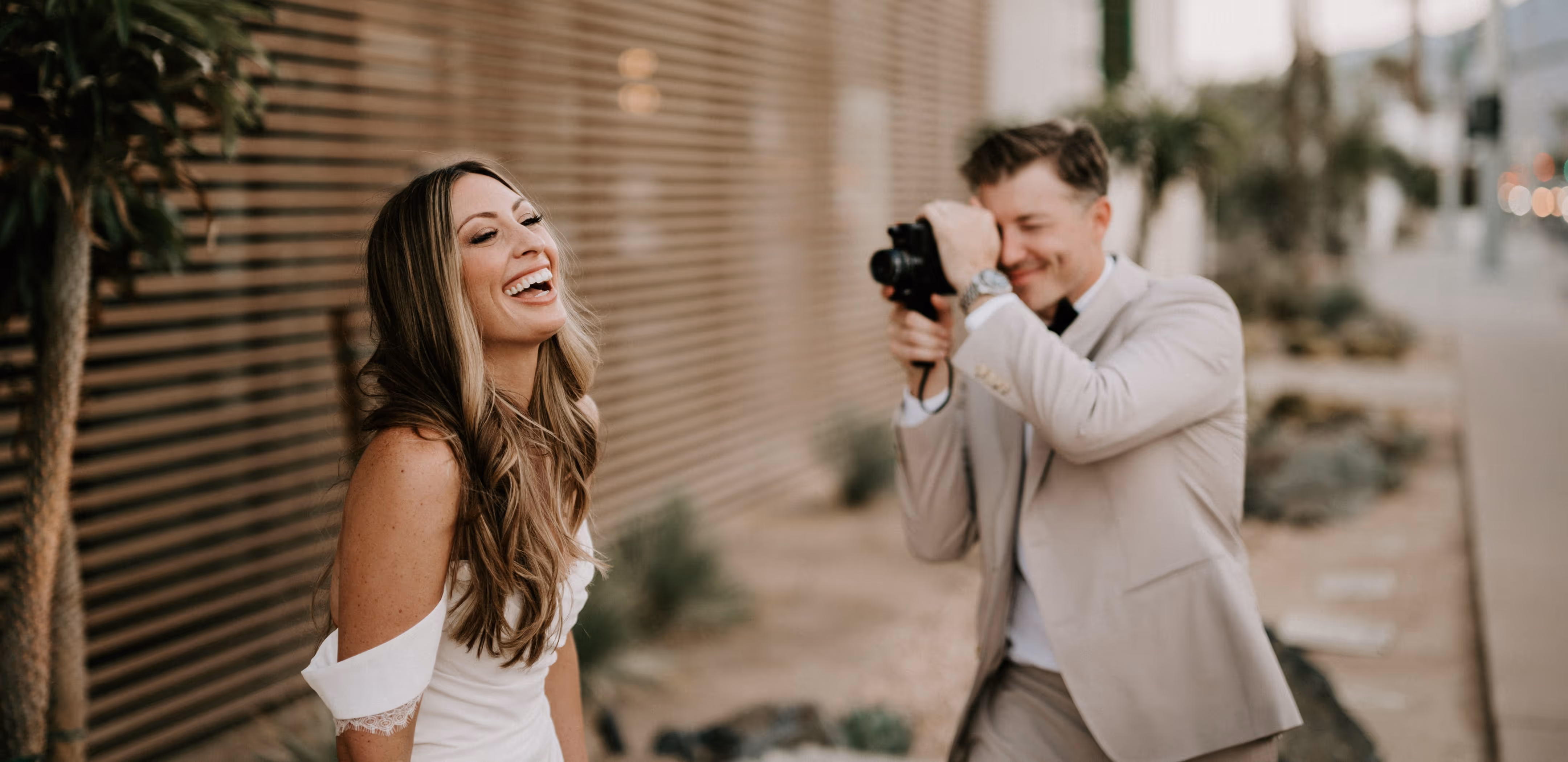 A smiling woman in a white off‑shoulder dress laughs against a slatted wooden wall as a man in a beige suit photographs her with a handheld camera outdoors.