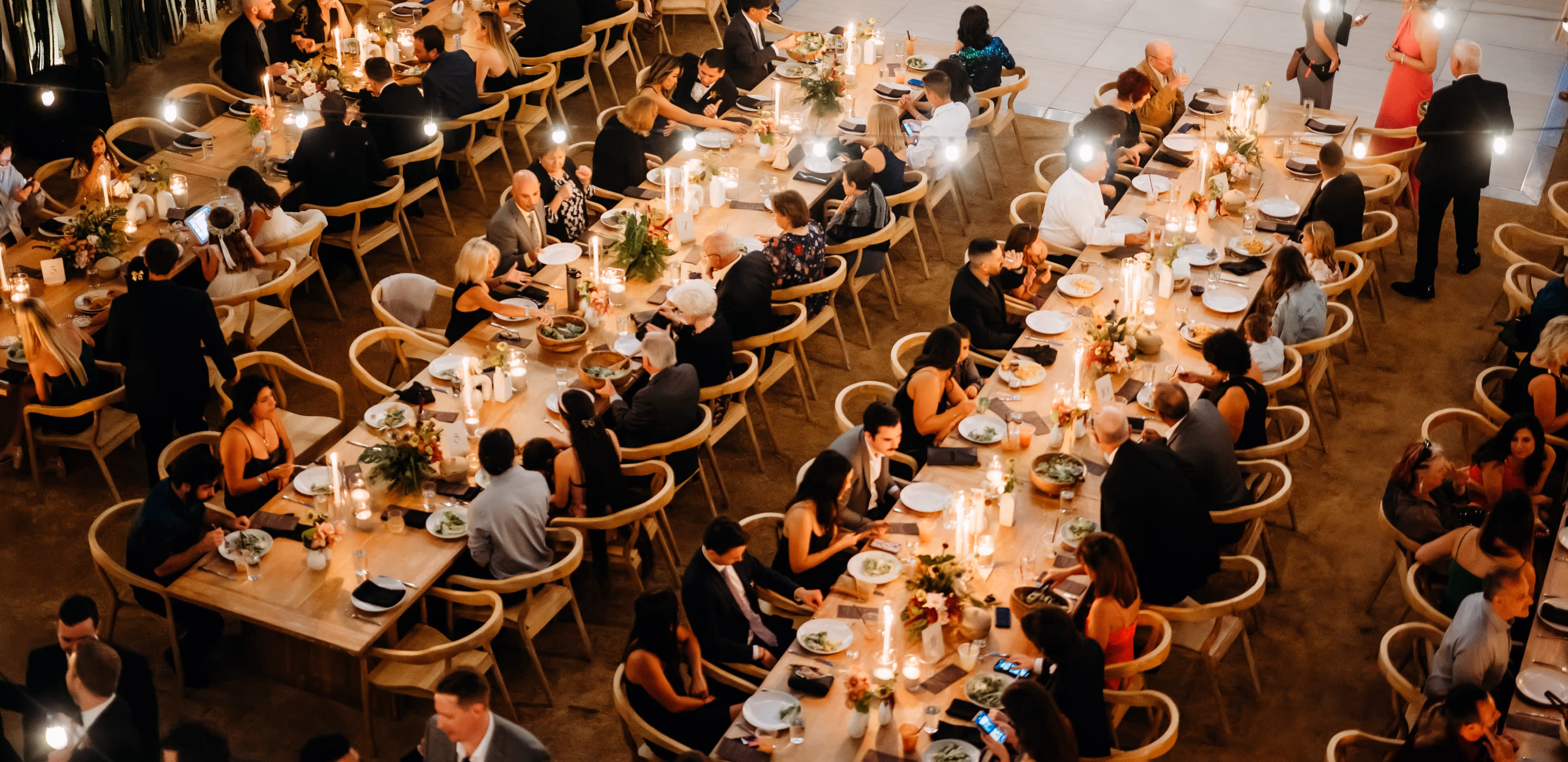 Overhead view of guests dining at long wooden banquet tables set with plates, salads and candles under warm string lights in a reception hall.