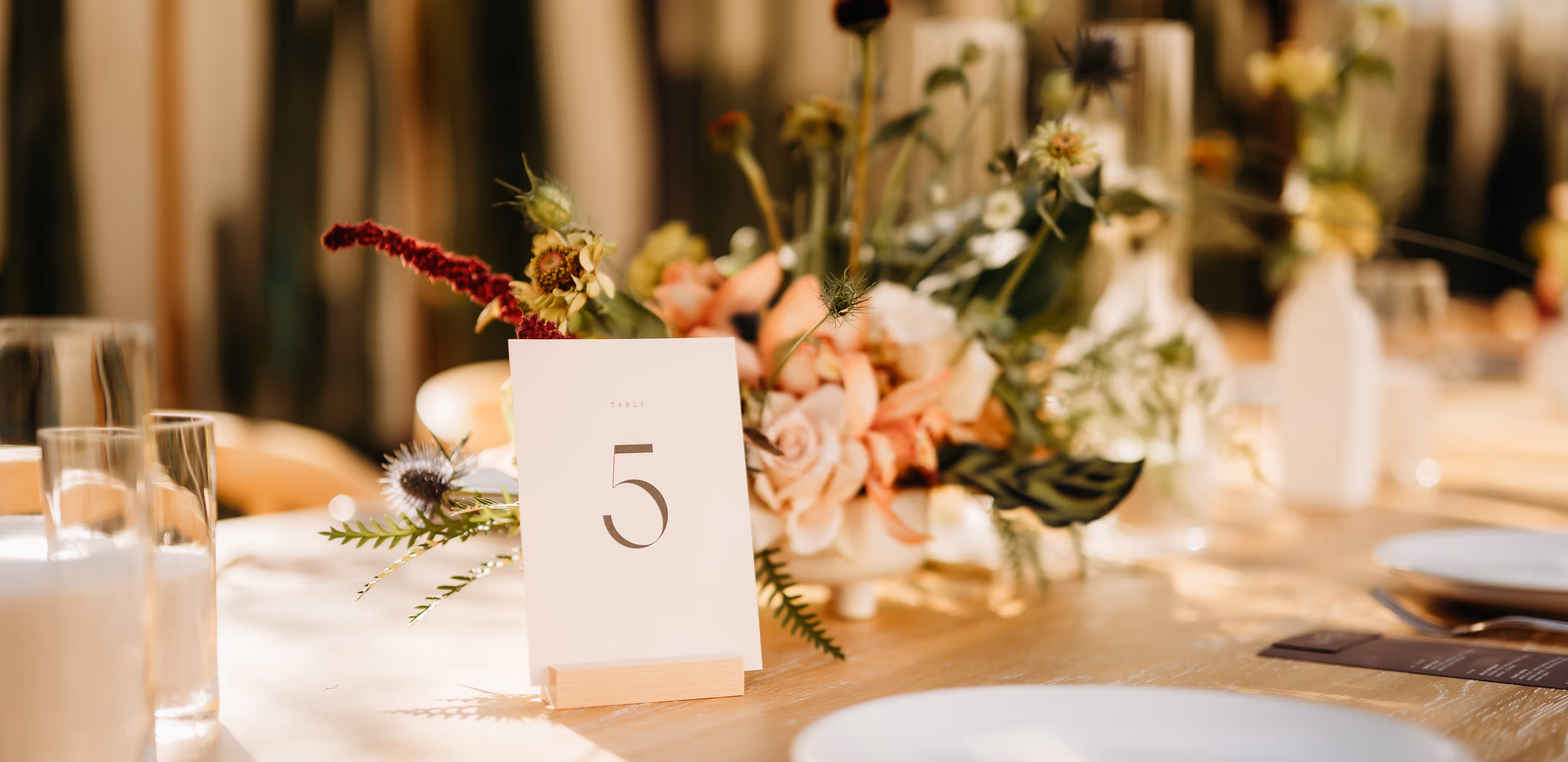 Close‑up of a wooden table centerpiece showing a “5” table number card in a stand, surrounded by pastel floral arrangements and empty glassware.
