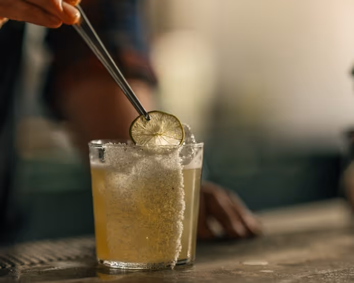 Close-up of a salt-rimmed margarita glass being garnished with a lime wheel held by metal tongs, set on a bar counter with a softly blurred background.