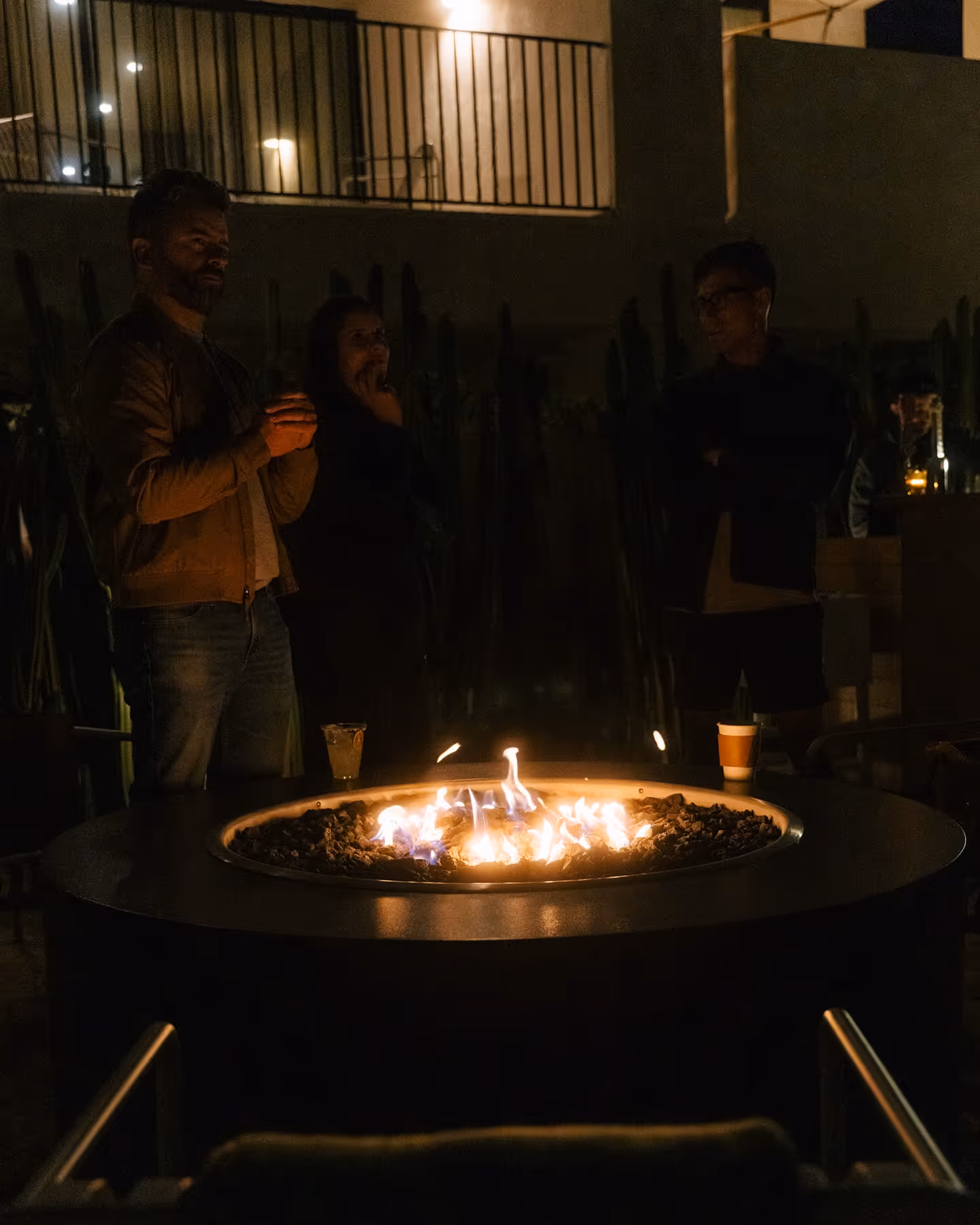 Three people stand around a circular outdoor fire pit at night, flames flickering over lava rocks. One holds a plastic cup, another a paper coffee cup, with tall plants and patio lights behind.