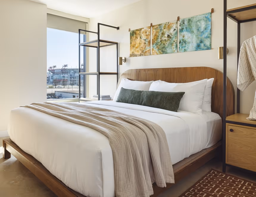 Modern bedroom suite featuring a low wooden platform bed with white linens, beige throw blanket and green accent pillow; industrial metal shelving, abstract wall art, and a large window with a stadium view.