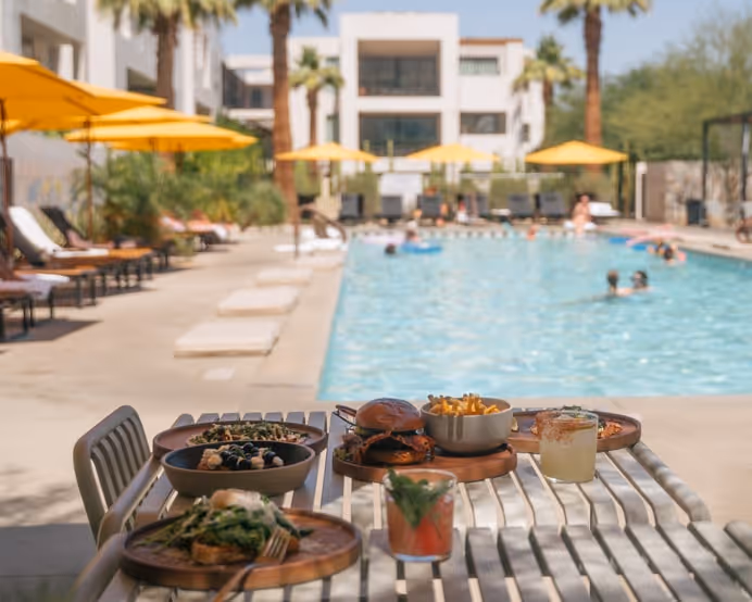 A sunny poolside table set with a burger, fries in a white bowl, two wooden bowls of salad, and colorful cocktails, with yellow umbrellas, sun loungers, and palm trees in the background.
