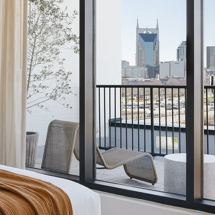 Modern hotel room with a balcony, lounge chairs, potted plant, and city skyline view featuring a distinctive skyscraper in the distance.