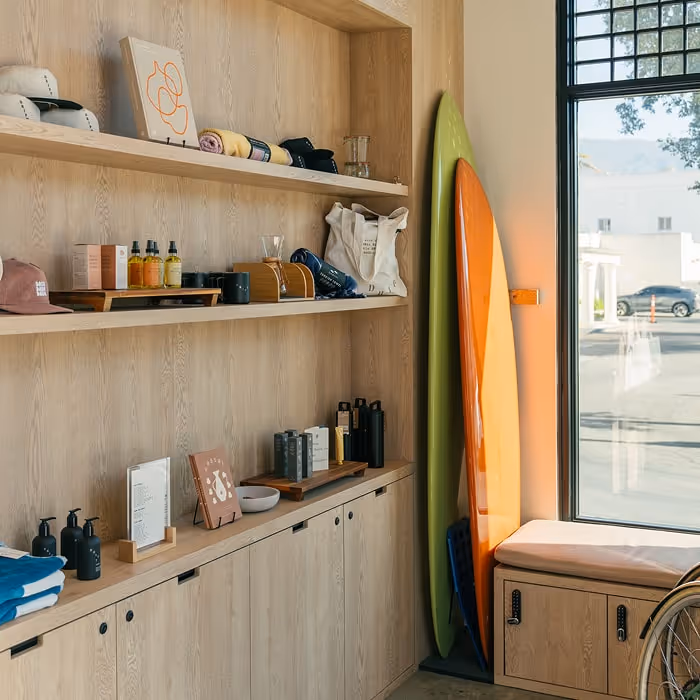 Surf shop corner with wooden shelves displaying apparel, skincare, and accessories, alongside two colorful surfboards by a sunny window.