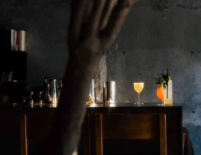 Dimly lit bar with wooden chairs, cocktail shakers, and three colorful drinks on the counter against a rustic gray wall.