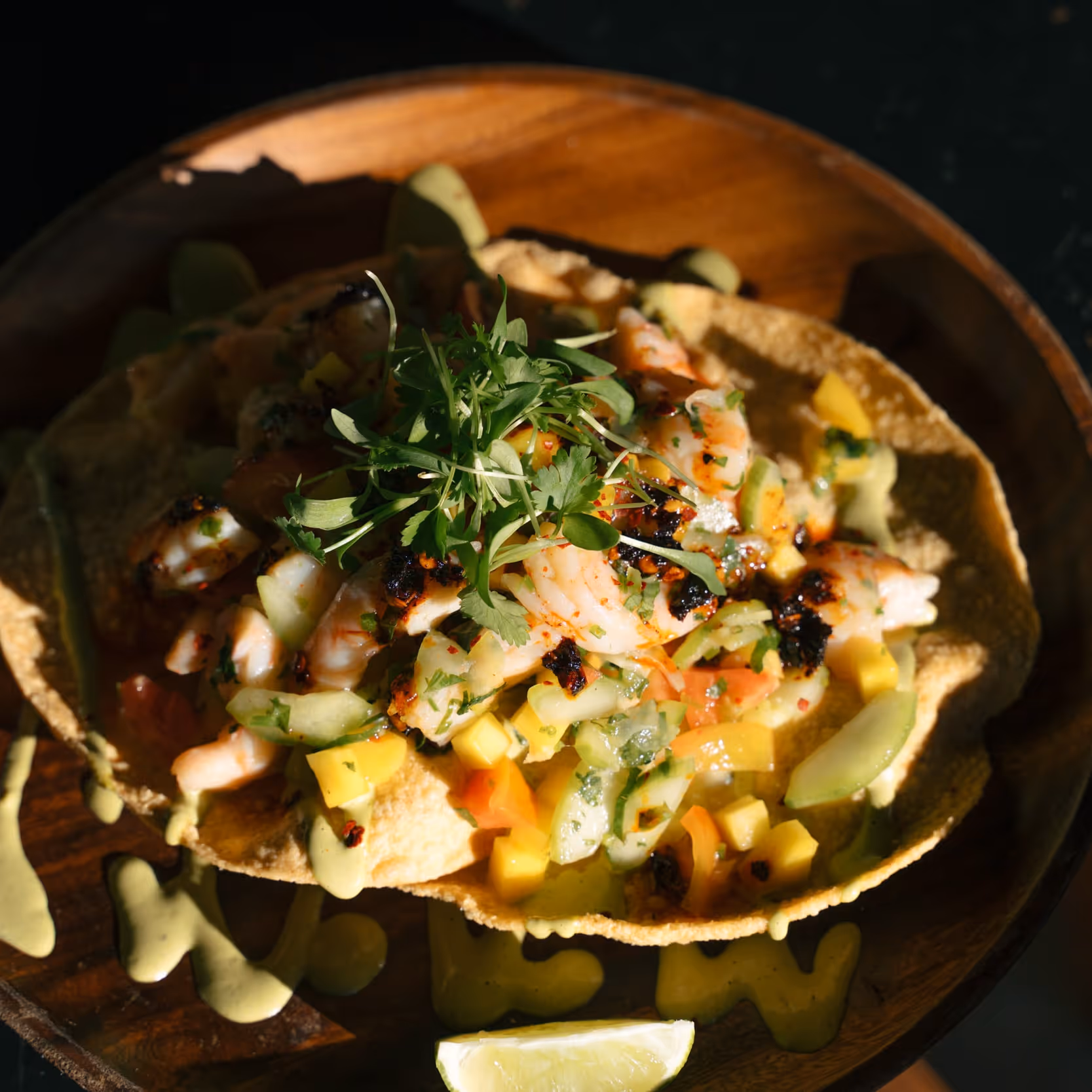 Shrimp tostada topped with cucumber, mango, herbs, and chili flakes, served on a wooden plate with lime.
