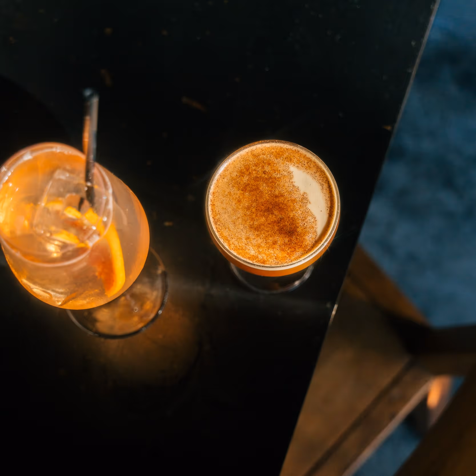 Overhead view of two cocktails on a dark table, one with an orange slice and ice, the other topped with foam and cinnamon.
