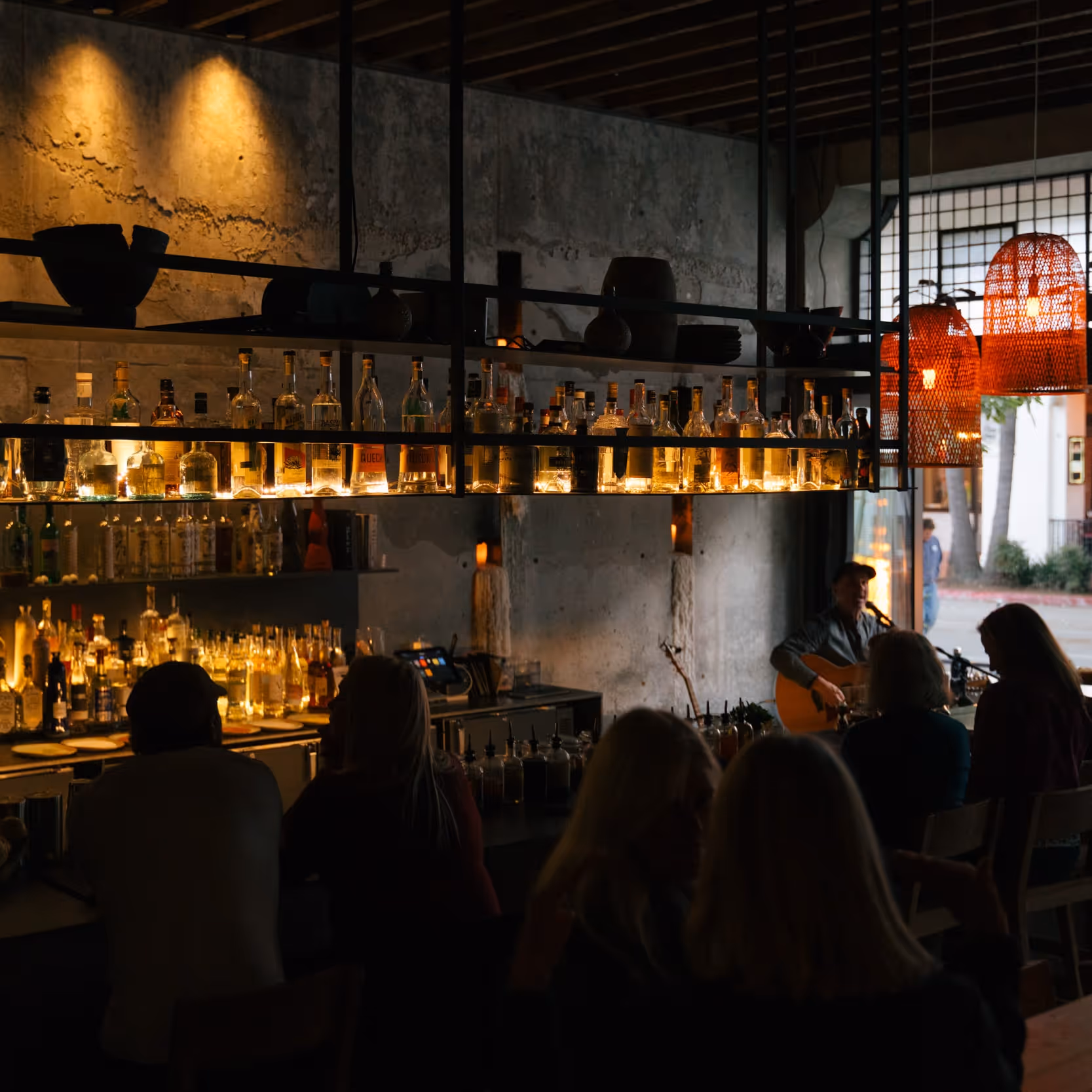Dimly lit bar with shelves of liquor bottles glowing in warm light, people seated at the counter, and a musician playing guitar in the corner.