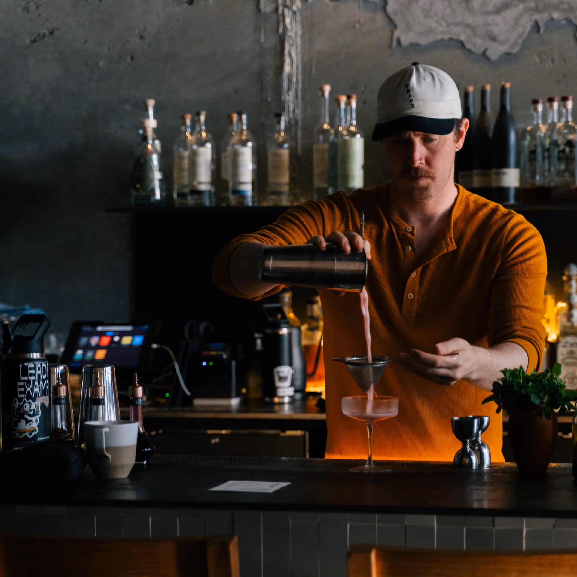 Bartender in an orange shirt pours a pink cocktail through a strainer into a glass at a rustic bar.