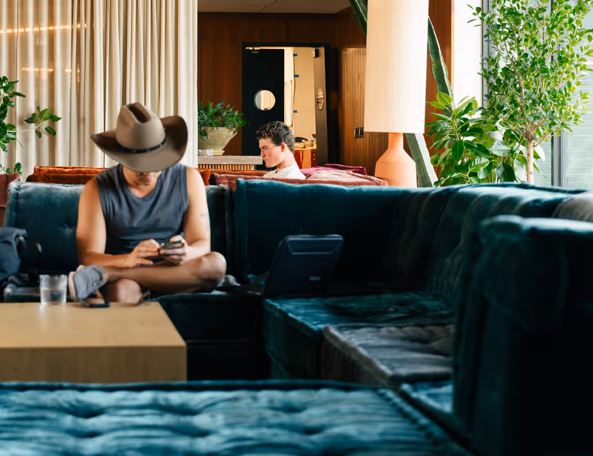 Man wearing a cowboy hat sitting cross-legged on a velvet sofa using his phone in a stylish modern lounge with plants and cozy seating.
