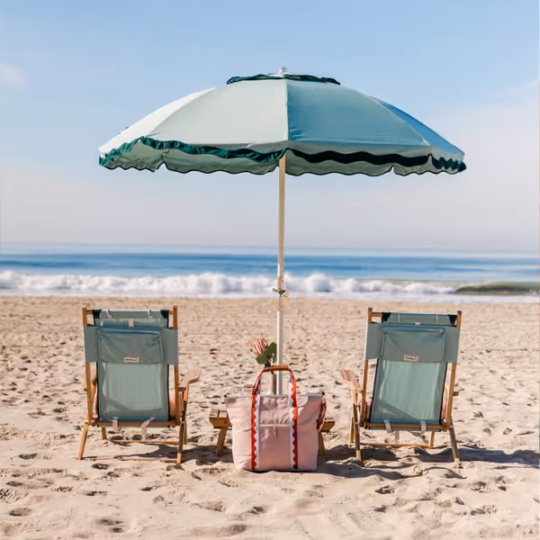 Two beach chairs and a turquoise umbrella with a tote bag set up on a sandy beach by the ocean waves