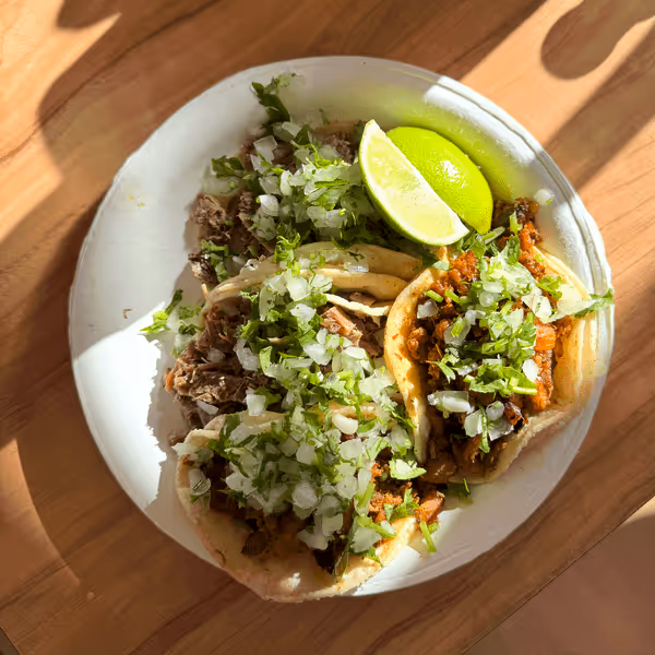 Authentic Mexican street tacos filled with seasoned meat, fresh cilantro, diced onions, and lime wedges served on a white plate in natural sunlight.