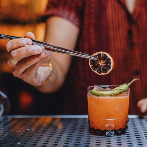 Bartender adding a dried citrus garnish to a bright orange cocktail at Pushing Daisies bar, showcasing craft mixology and vibrant nightlife atmosphere.