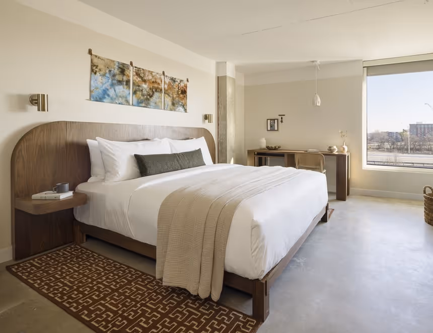 Spacious modern hotel room with king bed, wood furniture, neutral decor, large window, and work desk.