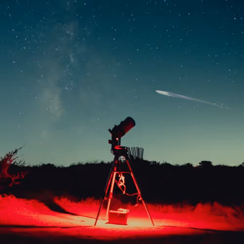 Telescope illuminated by red light under a clear night sky with stars and a visible comet, ideal scene for stargazing or astrophotography.