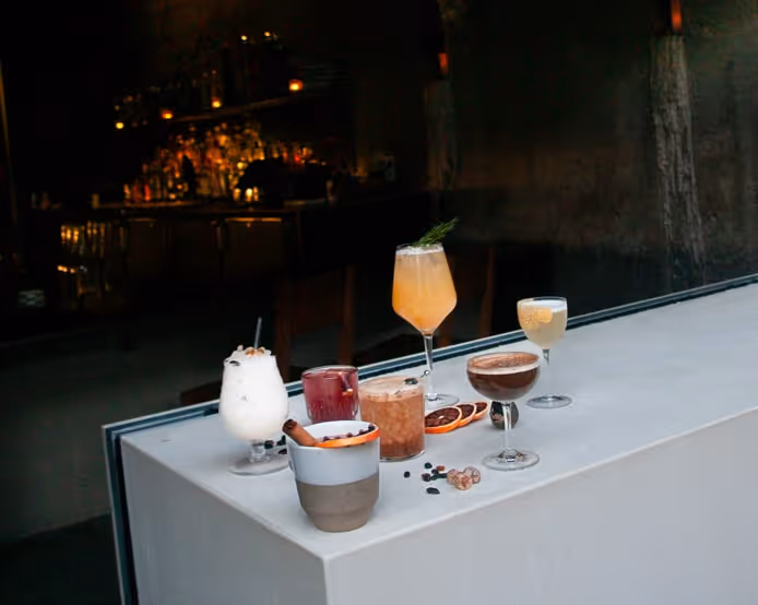 A minimalist bar counter with six colorful cocktails and drinks displayed under warm lighting, featuring garnishes of citrus, herbs, and spices at a modern dimly lit bar.