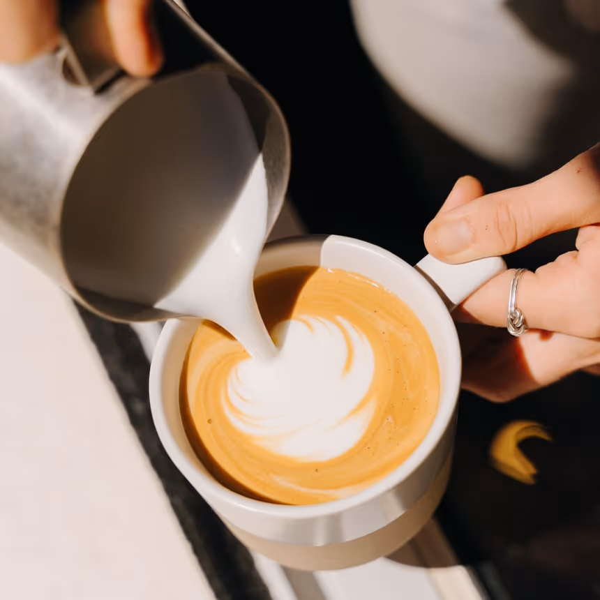 Close-up of barista pouring steamed milk to create latte art in a white cup of specialty coffee at a modern café.
