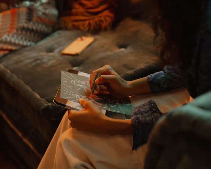 Artist sketching on a digital tablet while sitting on a sofa, creating colorful artwork in a softly lit creative space.