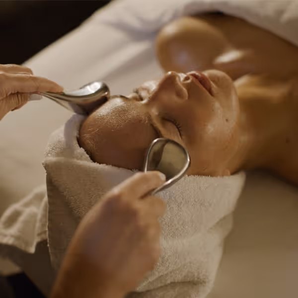Person receiving a relaxing facial treatment with cooling metal massage tools applied to the forehead, lying on a spa table with a towel wrapped around the head.