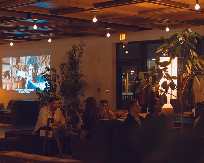 Cozy lounge event scene with guests chatting under string lights while a film plays on a projector screen.
