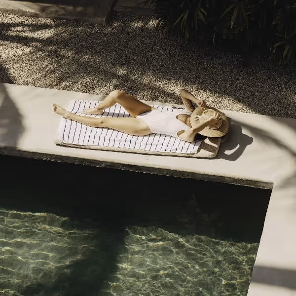 Woman relaxing poolside on a sun lounger, wearing a straw hat and swimsuit, enjoying a calm luxury resort atmosphere.