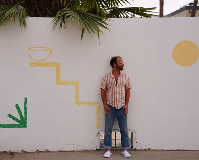 Man in striped linen shirt and jeans standing against white wall with minimalist yellow staircase and sun mural