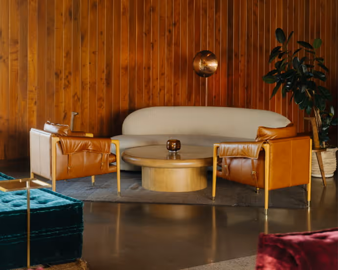 Mid-century modern living room with tan leather chairs, curved white sofa, round wood coffee table, and warm wood paneling