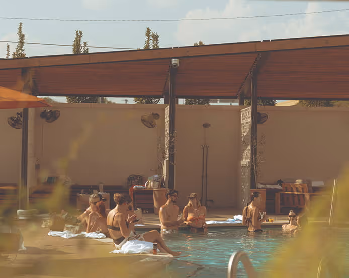 People relaxing poolside at a modern outdoor pool lounge with wooden pergola, string lights, and industrial fans.