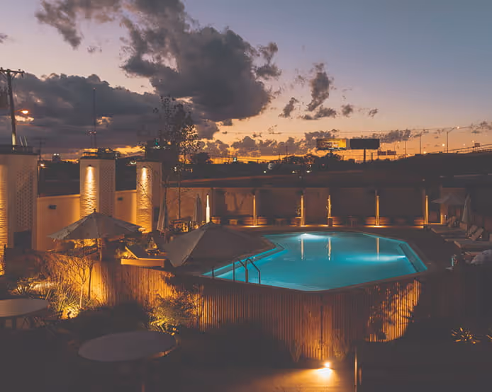 Illuminated turquoise swimming pool on a rooftop deck at twilight with lounge chairs, umbrellas, and a dramatic sunset sky.