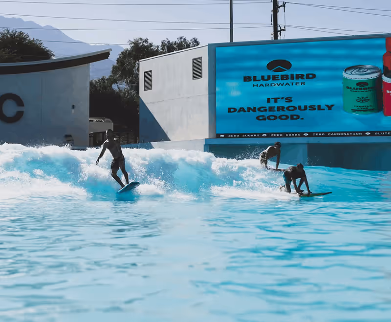 Surfers riding artificial waves at Palm Springs Surf Club with Bluebird Hardwater advertisement on large screen