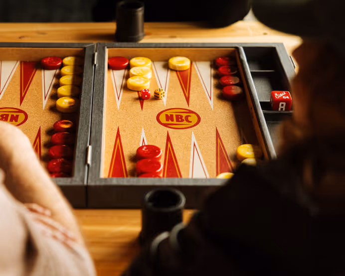A vintage NBC-branded backgammon set with red and yellow checkers mid-game, featuring dice and leather cup on wooden table.