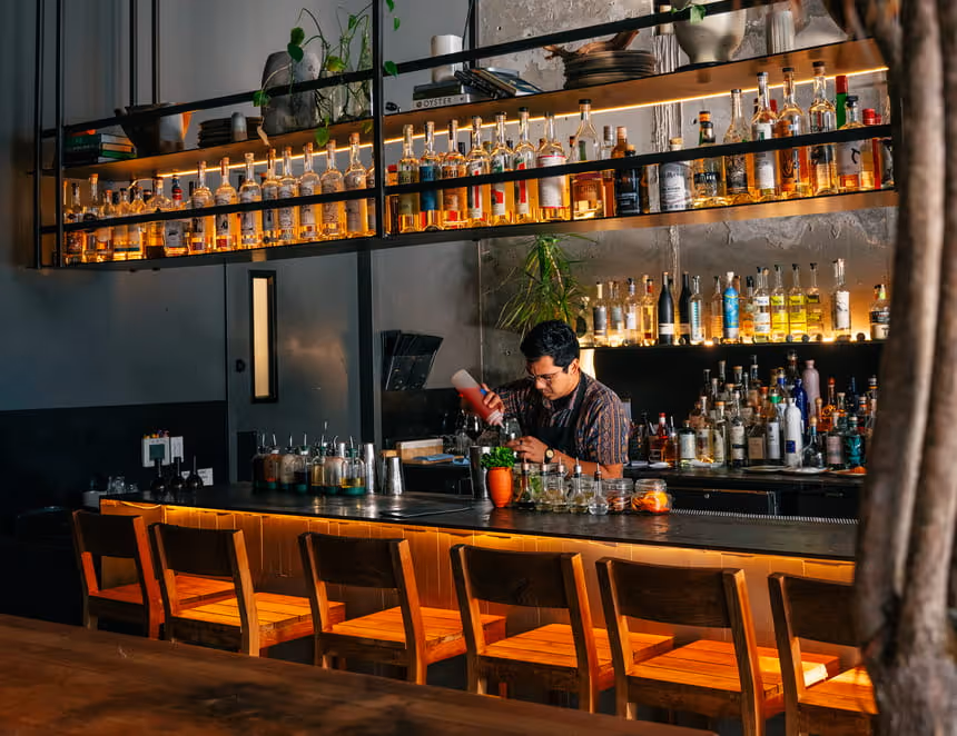 A bartender prepares craft cocktails behind a sleek dark bar lined with wooden stools and illuminated shelves of spirits.