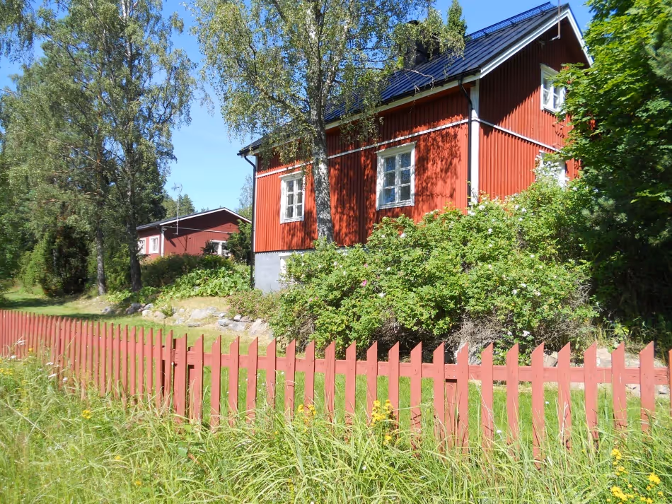 Red wooden house with white-trimmed windows behind a red picket fence surrounded by green bushes and trees under a clear blue sky.