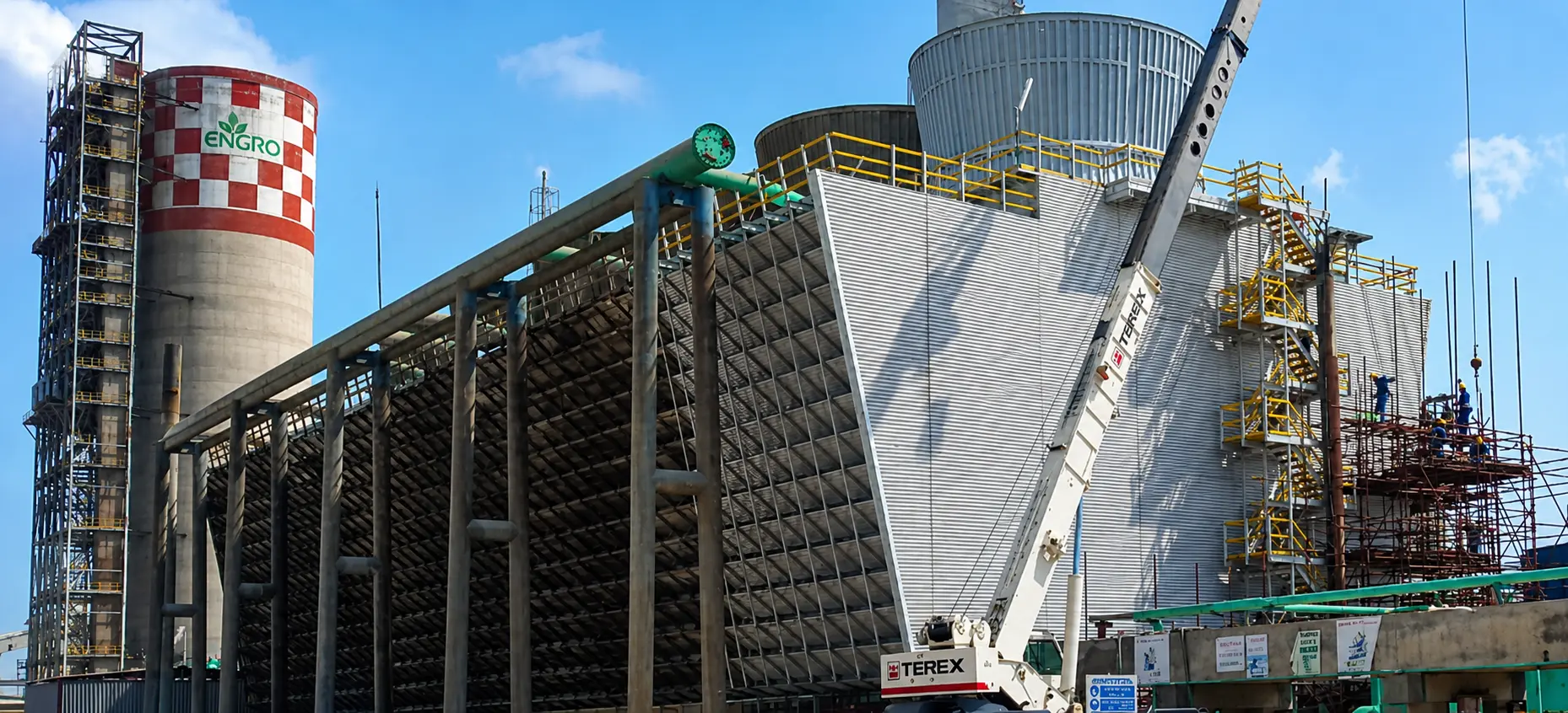 Industrial facility with large Engro-branded tower, cooling tower structures, scaffolding, and a Terex crane lifting materials.