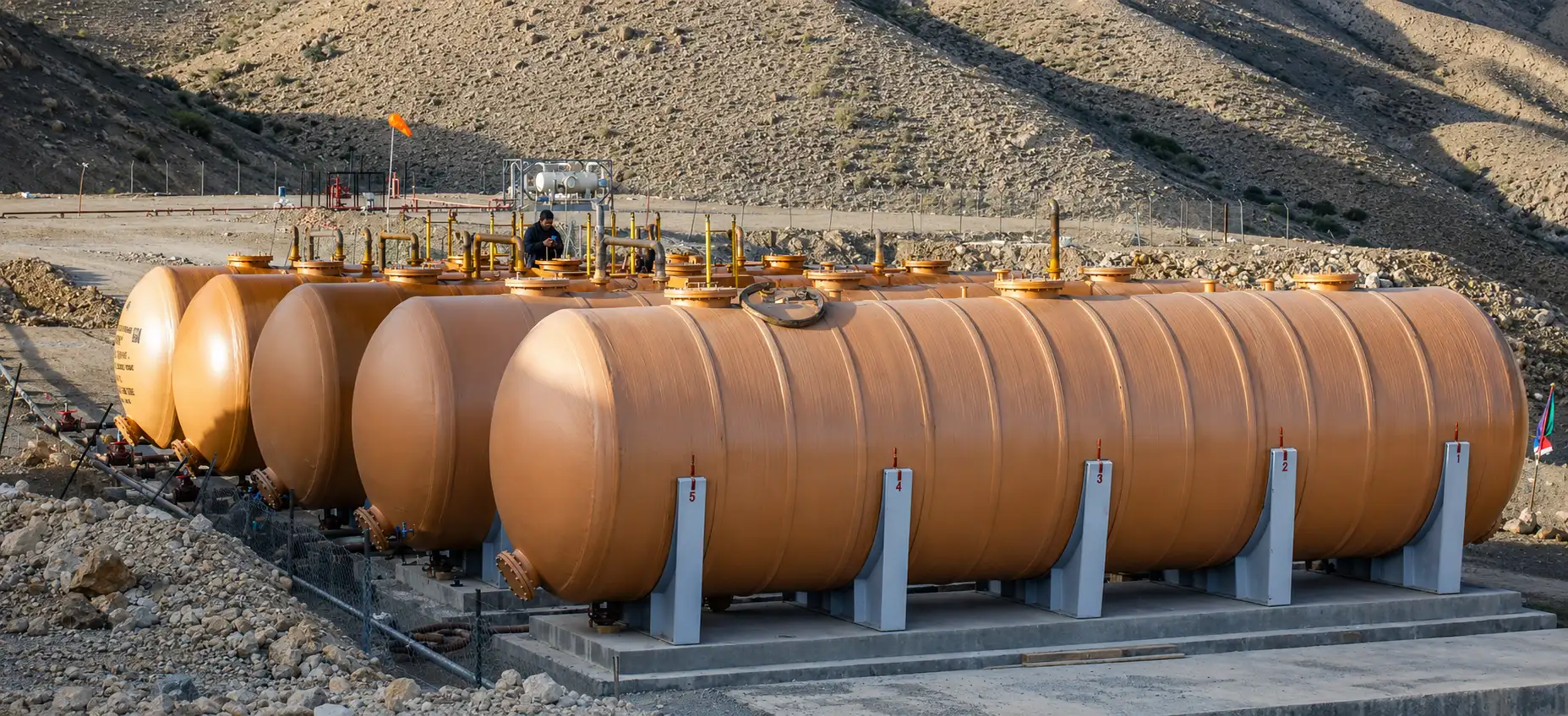 Large orange industrial storage tanks lined up outdoors on a concrete platform in a desert-like area with hills in the background.