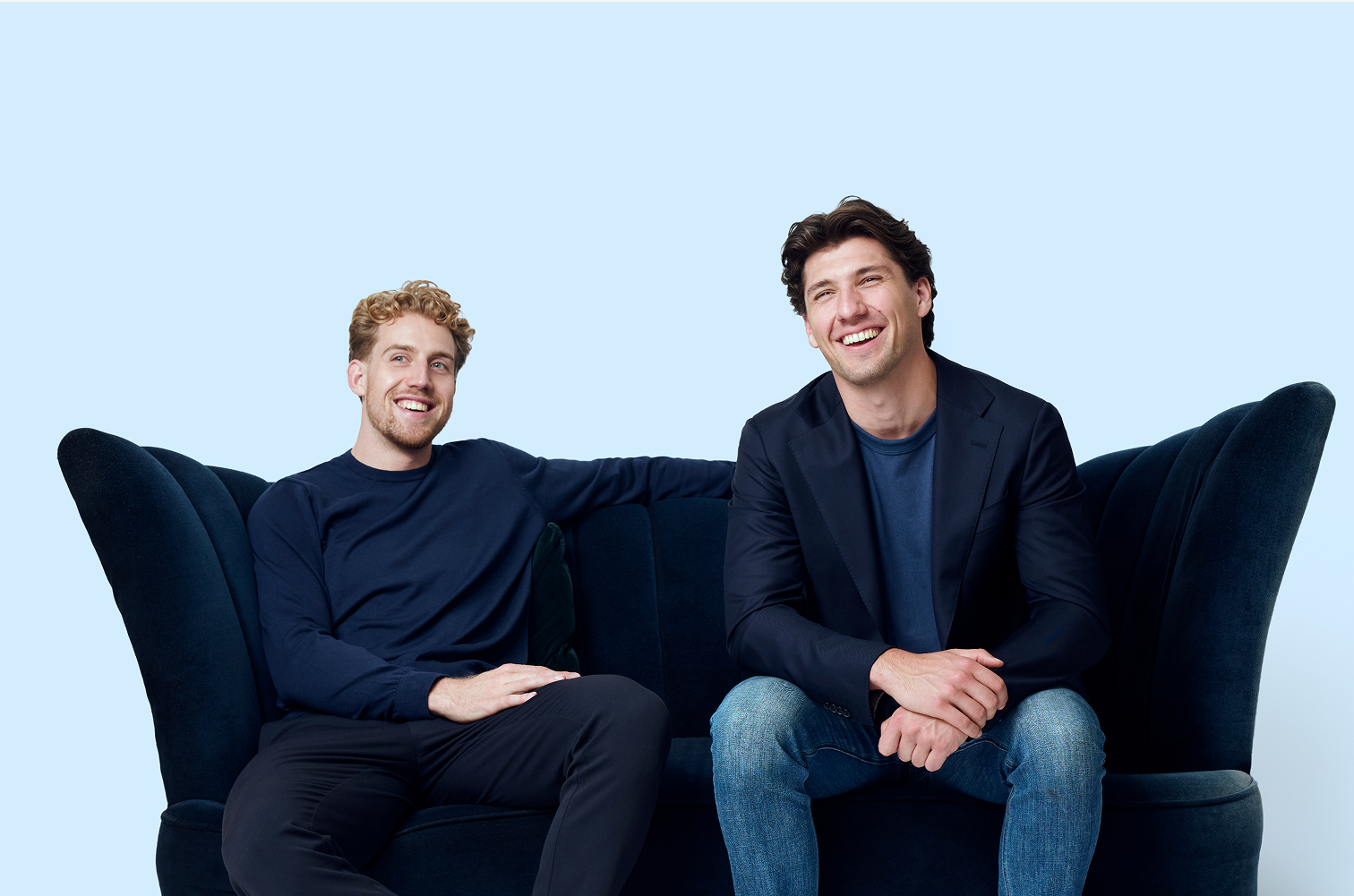 Twee lachende mannen zitten ontspannen op een donkerblauwe bank tegen een lichte achtergrond.