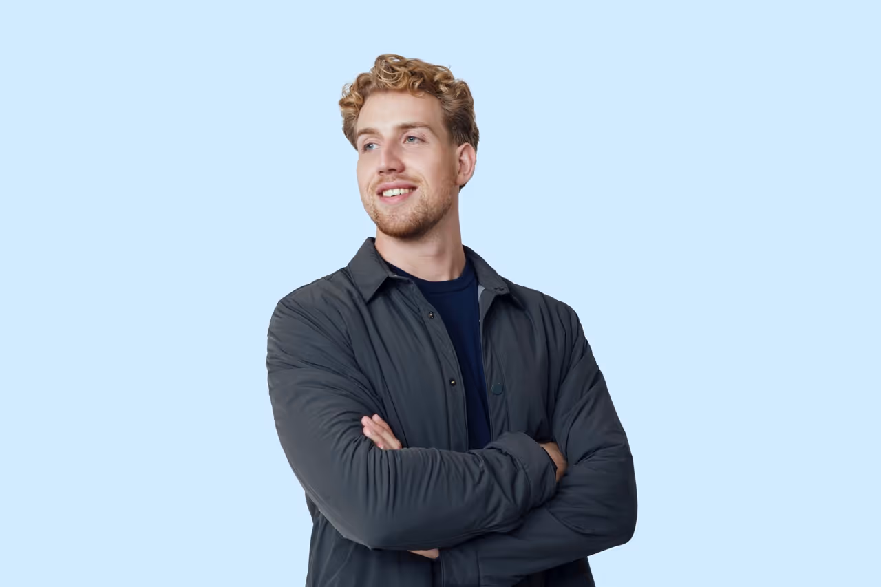 Jonge man met gekruld blond haar in een donkergrijze jas lacht en kijkt naar links, met gekruiste armen tegen een lichtblauwe achtergrond.